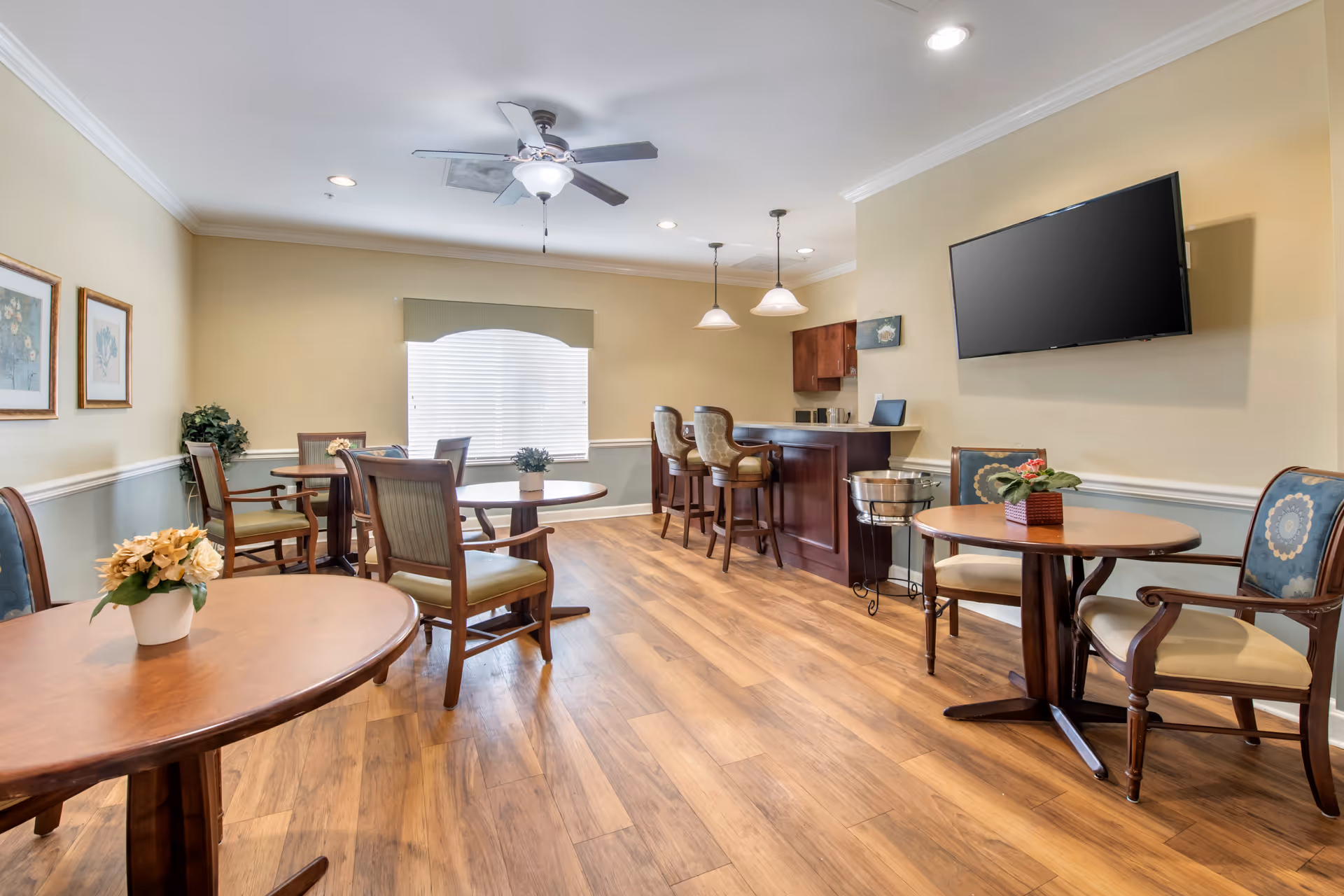 A bright and spacious common area with wooden floors, several round tables with chairs, a ceiling fan, a wall-mounted flat-screen TV, and a small bar counter with bar stools. The room has beige walls with white trim and a large window with blinds letting in natural light.