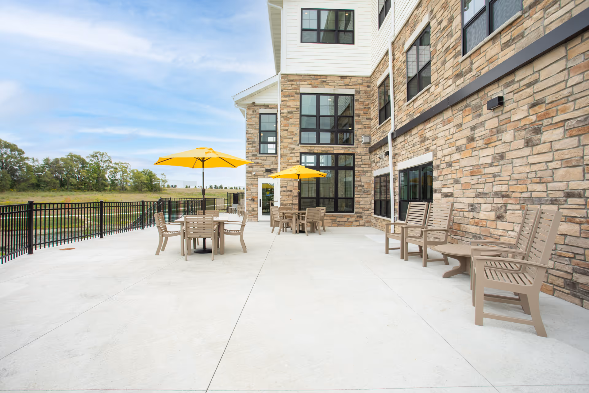 Outdoor patio area at Holland Farms Senior Living with beige chairs and tables, two yellow umbrellas, a stone building facade, and a black metal fence overlooking a grassy field and trees under a partly cloudy sky.