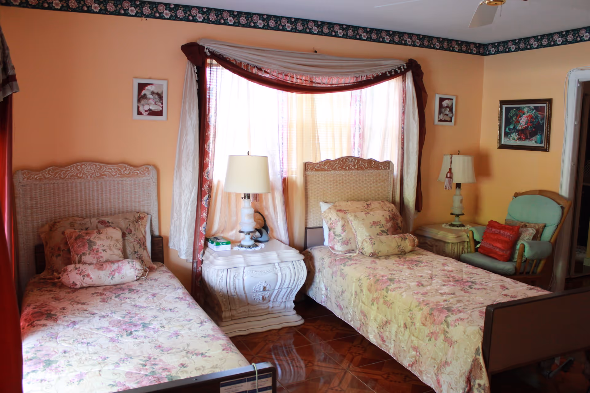 A cozy bedroom with two single beds featuring floral bedspreads and matching pillows. Between the beds is a white ornate nightstand with a lamp and some items on it. The room has peach-colored walls with floral border wallpaper near the ceiling, framed floral artwork, a window with sheer curtains and burgundy drapes, and a cushioned rocking chair with red and floral pillows.