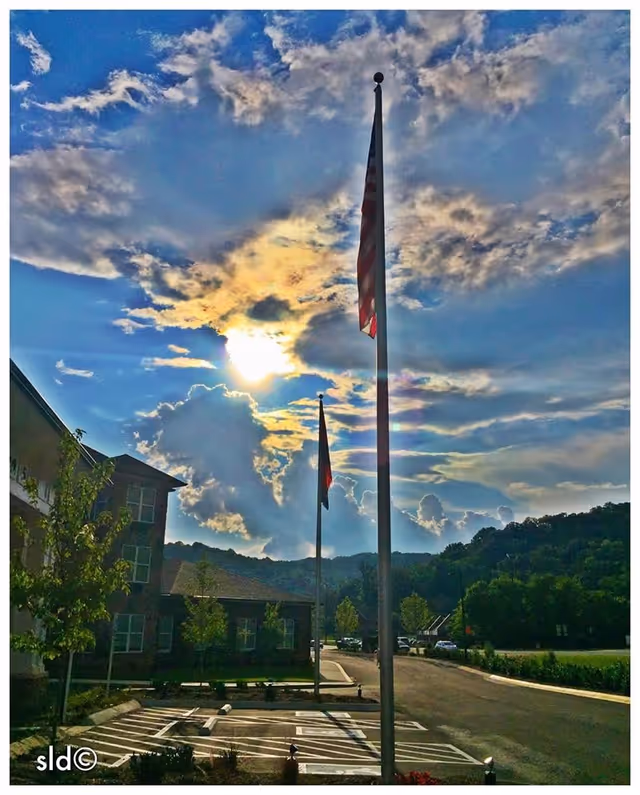 Outdoor view of Canterfield Senior Living facility parking area with two flagpoles displaying flags under a partly cloudy sky with the sun shining through. Trees, hills, and parts of the building are visible in the background.