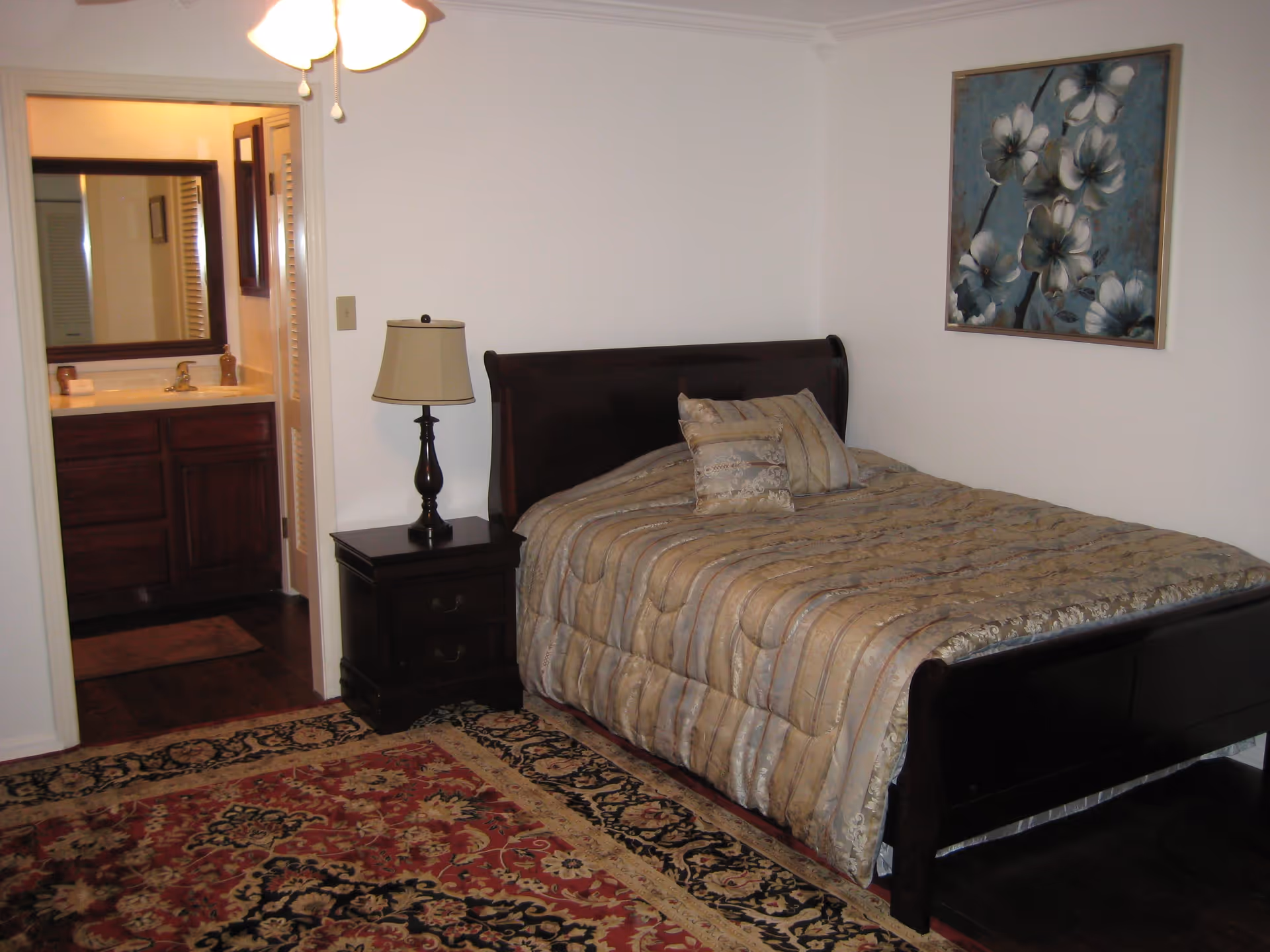 A bedroom with a dark wooden bed frame and a beige patterned comforter with matching pillows. There is a dark wooden nightstand with a lamp beside the bed. A floral painting hangs on the white wall above the bed. The room has a patterned area rug on the floor. An open doorway leads to a bathroom with a sink, mirror, and wooden cabinetry.