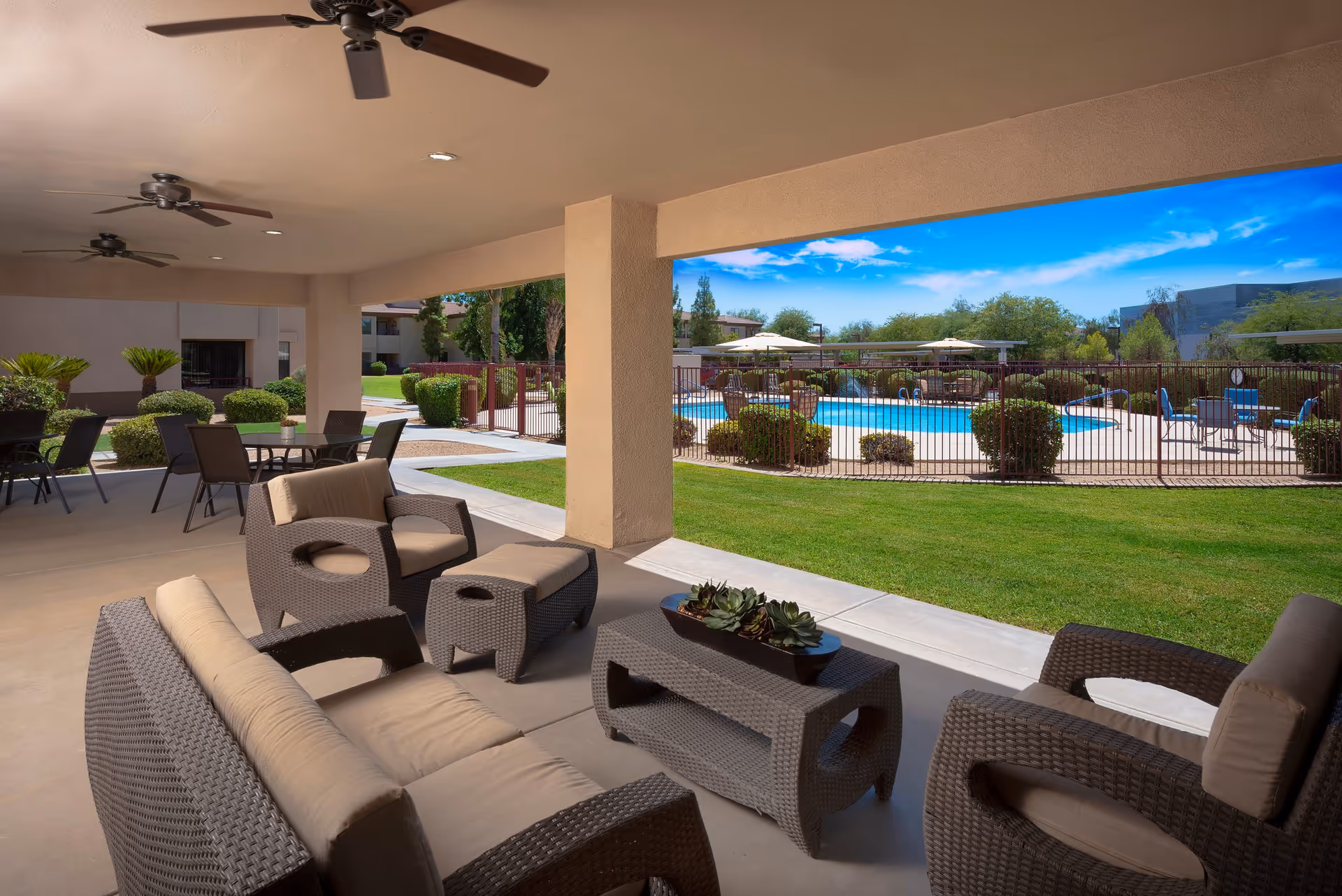 Covered outdoor patio area with wicker-style cushioned seating including a sofa, armchairs, ottoman, and a coffee table with a succulent centerpiece. Ceiling fans are mounted on the patio ceiling. Beyond the patio is a fenced swimming pool area with tables, chairs, and umbrellas, surrounded by green grass and shrubs under a bright blue sky.