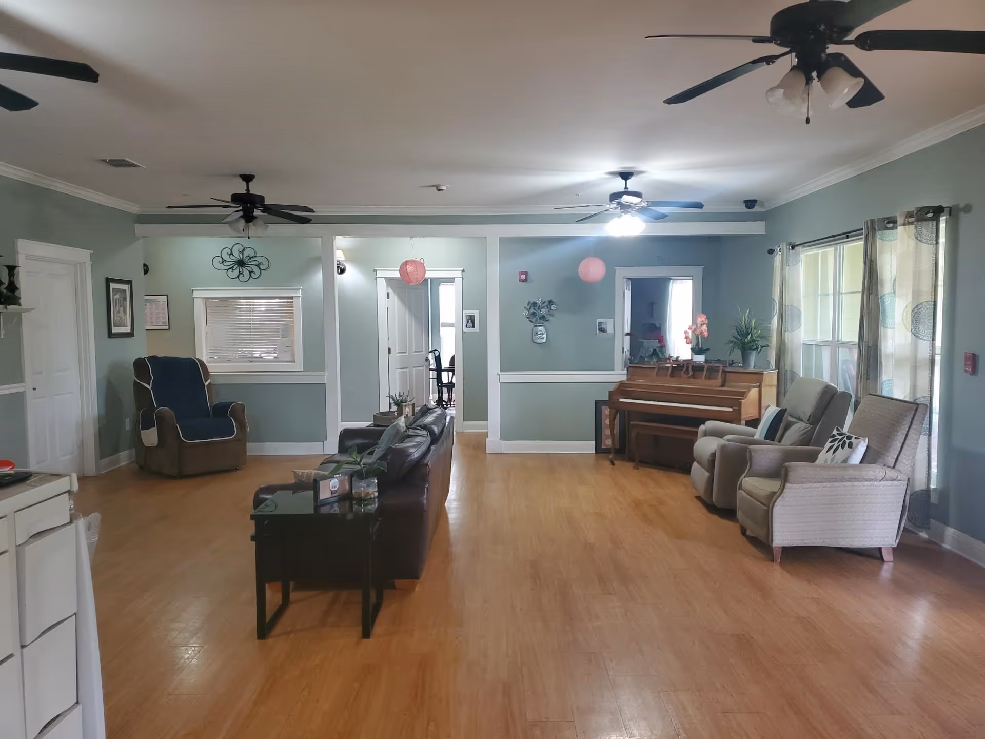 A spacious living room area with light green walls and wooden flooring. The room features a brown leather sofa, a side table with plants, two beige armchairs, a piano with decorative items on top, and a recliner chair with a blue cover. Ceiling fans with lights hang from the ceiling, and large windows with patterned curtains allow natural light to enter the room.