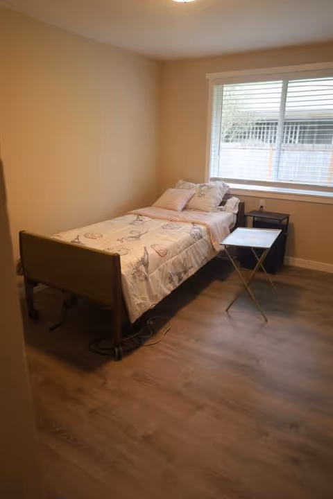 A small bedroom with a single bed against the left wall, covered with a light-colored bedspread featuring a pattern. There are several pillows on the bed. Next to the bed is a small black nightstand and a folding tray table. A large window with white blinds is on the right wall, letting in natural light. The floor is wood, and the walls are painted a light beige color.