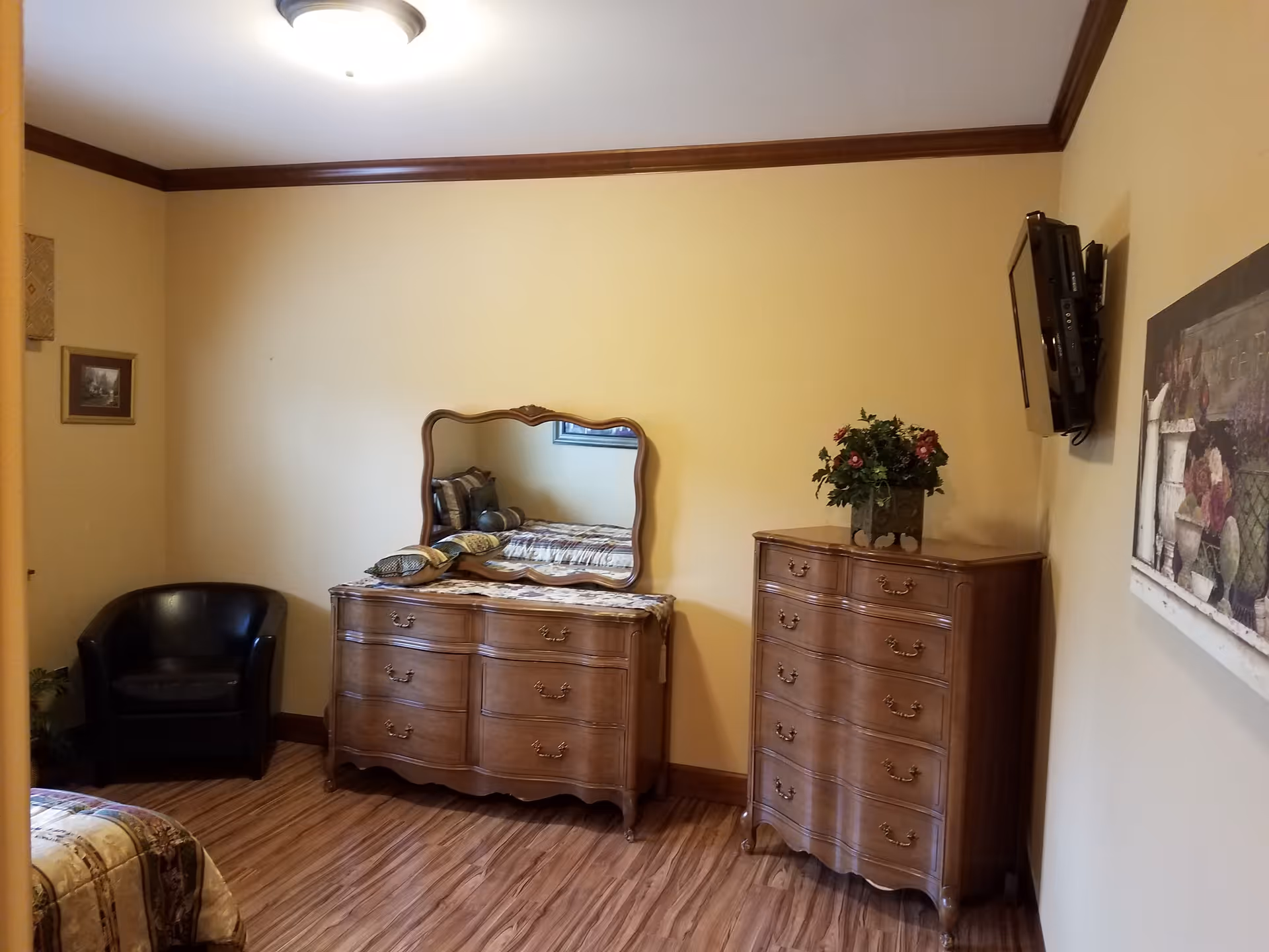Bedroom with two wooden dressers and mirror, a wall-mounted TV, a black armchair, and hardwood floor.