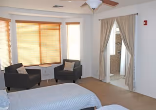 A bright bedroom with two beds covered in light blue bedding, two dark armchairs with pillows, a small round table between the chairs, large windows with wooden blinds, and an open doorway with beige curtains leading to a bathroom.
