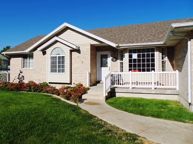 Exterior view of a single-story brick building with a sloped roof, white framed windows, a white front door, and a small porch with white railings. There is a concrete walkway leading up to the entrance, surrounded by green grass and flower beds with red and pink flowers. The sky is clear and blue.