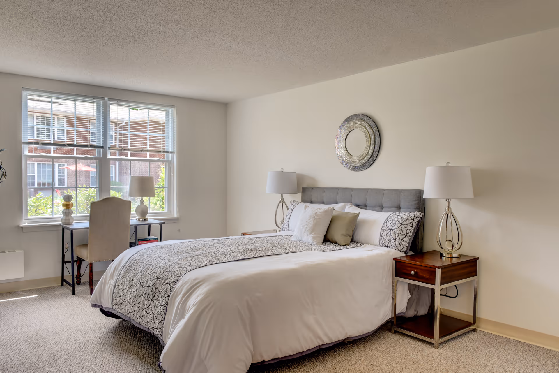 Well-lit bedroom with a made bed, bedside tables and lamps, a decorative mirror above the headboard, and a small desk by the window.