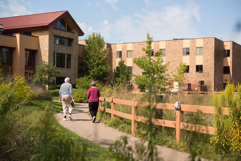 Two older adults walk along a landscaped path beside a wooden fence toward a brick senior living building.
