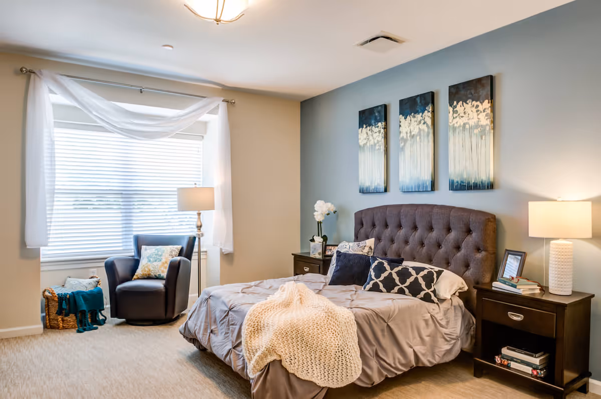 A cozy bedroom with a large bed featuring a tufted headboard, multiple decorative pillows, and a knitted throw blanket. On either side of the bed are dark wooden nightstands with lamps, books, and a framed photo. A comfortable armchair with a decorative pillow sits near a window with white blinds and sheer curtains. The walls are painted in soft beige and blue tones, with three abstract paintings hanging above the bed.