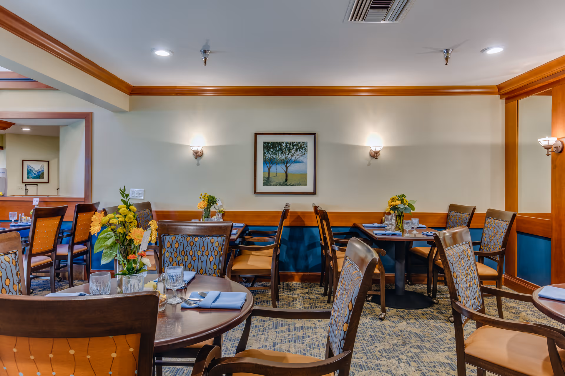 A dining room in a senior living facility with several wooden tables and chairs arranged neatly. Each table has place settings with napkins, glasses, and small flower arrangements. The walls are light-colored with wooden trim and sconces providing soft lighting. A framed picture of trees hangs on the wall.