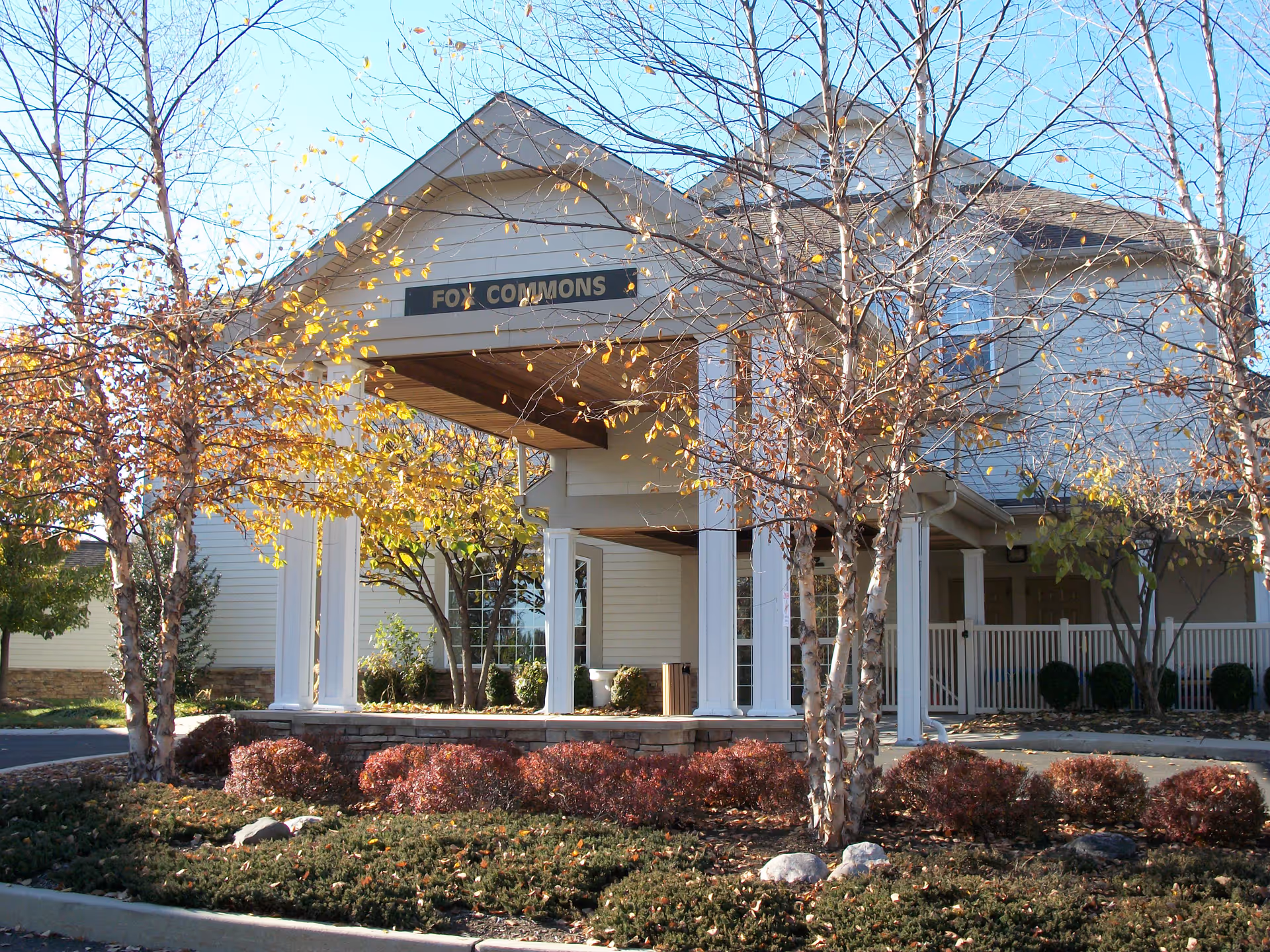Exterior view of a building entrance with a covered porch supported by white columns. The sign above the entrance reads 'FOX COMMONS'. There are trees with autumn leaves and landscaped bushes in front of the building under a clear blue sky.