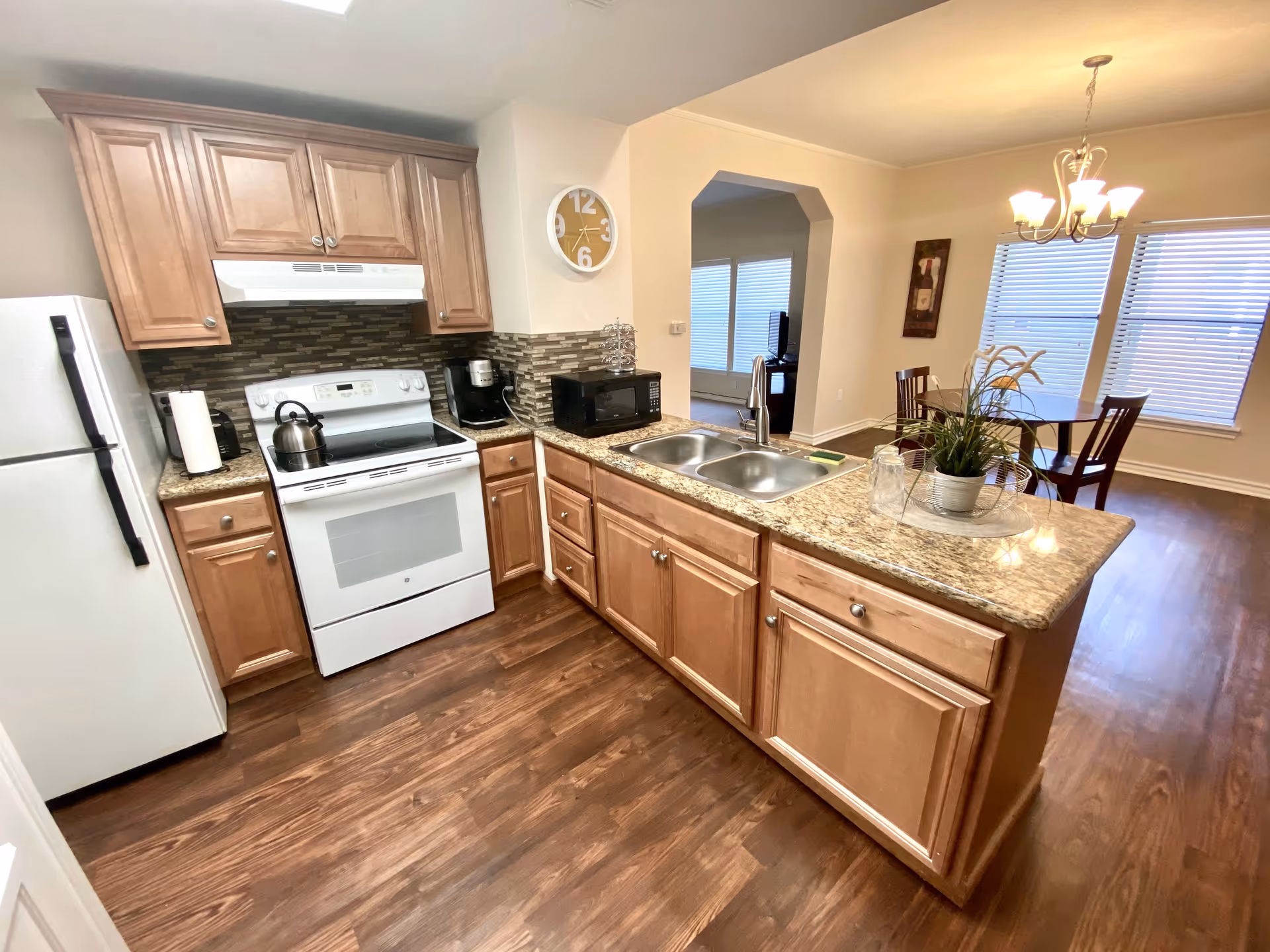 A modern kitchen with wooden cabinets, a white refrigerator, a white stove with a kettle on it, a microwave, and a double sink on a granite countertop. The kitchen opens into a dining area with a round wooden table, four chairs, a chandelier, and two large windows with blinds.