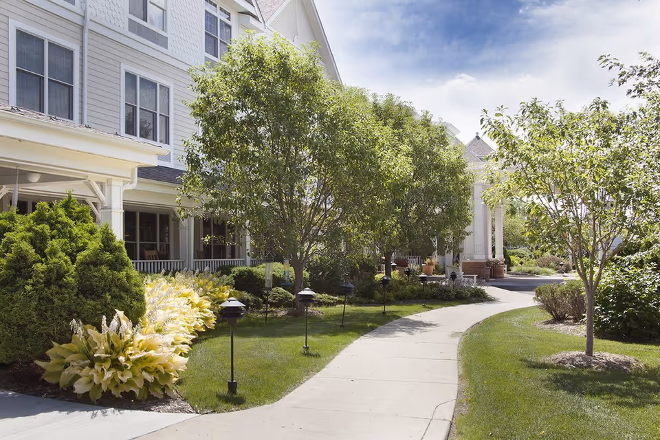 Curving concrete walkway flanked by manicured lawns, trees, and shrubs leading to the front entrance of a multi-story senior living building.