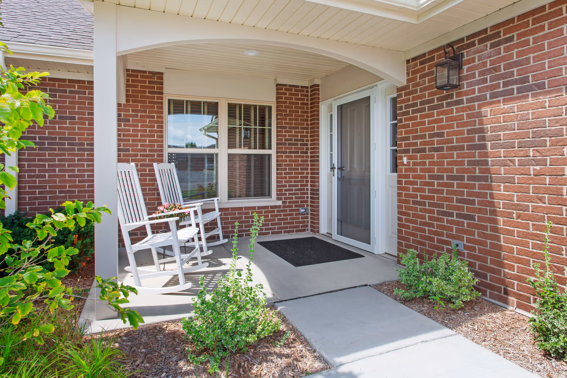 Front porch area of a brick building with two white rocking chairs, a small table with flowers, a black doormat, and some green shrubs and plants around the concrete walkway.