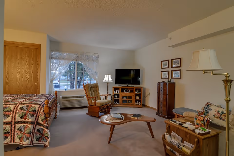 A cozy senior living room and bedroom area with a wooden bed covered with a quilt, a cushioned rocking chair, a wooden coffee table with books, a TV on a wooden stand, a tall chest of drawers, a floral-patterned sofa, and a floor lamp. The room has a window with sheer curtains letting in natural light.