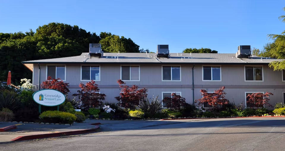 Exterior view of a two-story senior living facility building with multiple windows, surrounded by landscaped bushes and small red-leafed trees under a clear blue sky. A sign in front reads 'Greenridge Senior Care'.