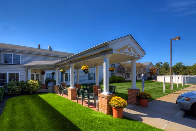 Outdoor covered seating area with green chairs and hanging flower pots in front of a white residential building under a clear blue sky. There are potted yellow flowers on brick pillars and a well-maintained lawn surrounding the area.