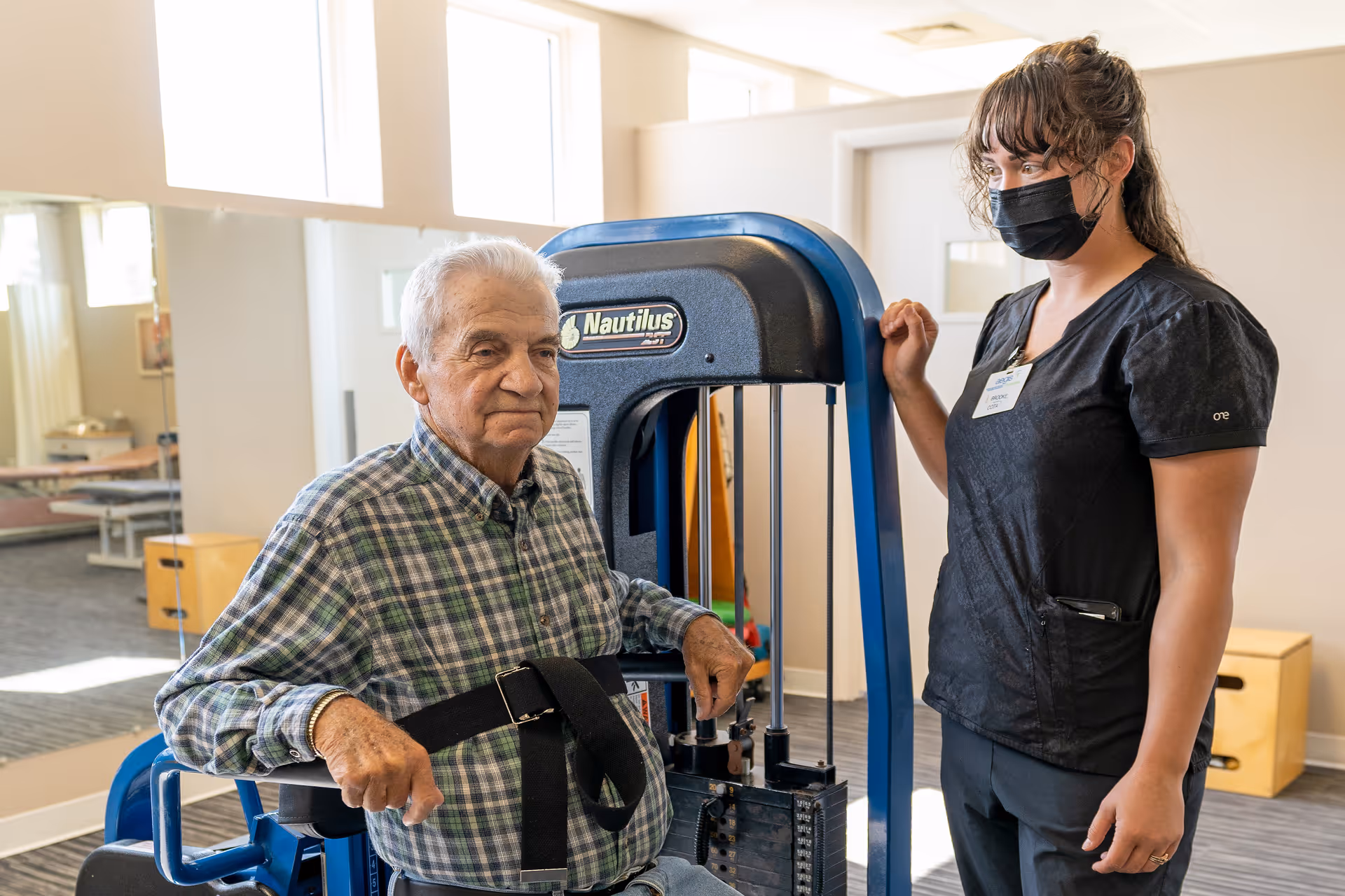 An elderly man using a Nautilus exercise machine in a rehabilitation center, assisted by a female staff member wearing a black mask and black scrubs. The room is well-lit with large windows and has exercise equipment in the background.