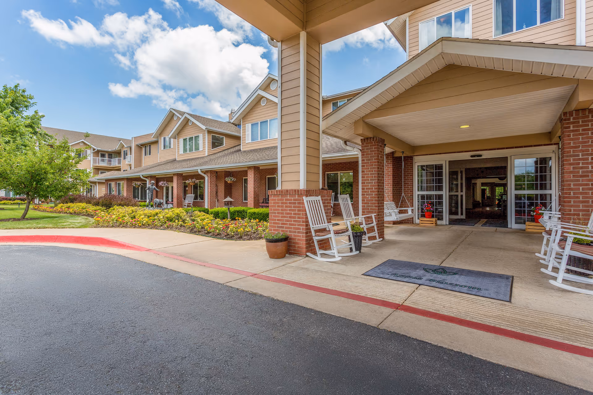 Exterior view of Morada Rogers senior living facility showing the entrance with a covered driveway, white rocking chairs, potted plants, and a well-maintained garden with flowers and shrubs under a partly cloudy sky.