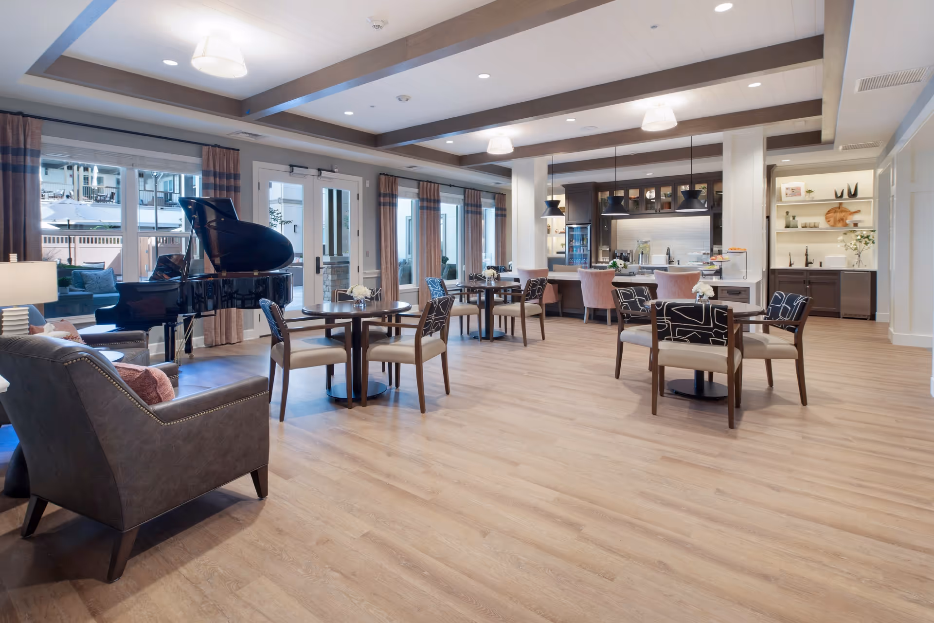 A spacious, well-lit common area with wooden flooring featuring several round tables with chairs, a black grand piano near large windows with curtains, comfortable armchairs, and a kitchen area with a counter and bar stools in the background.
