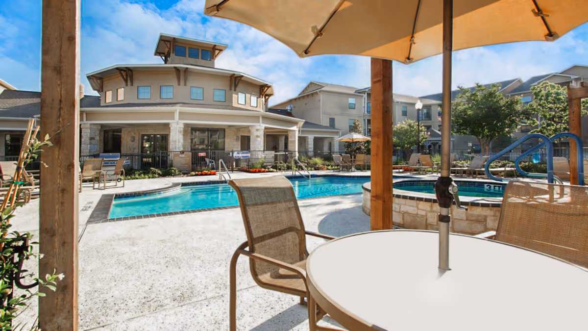 Outdoor pool area at Chateau McKinney with a round table and chairs under a large umbrella in the foreground. The pool is surrounded by lounge chairs, umbrellas, and landscaped greenery with a multi-story building in the background under a blue sky.