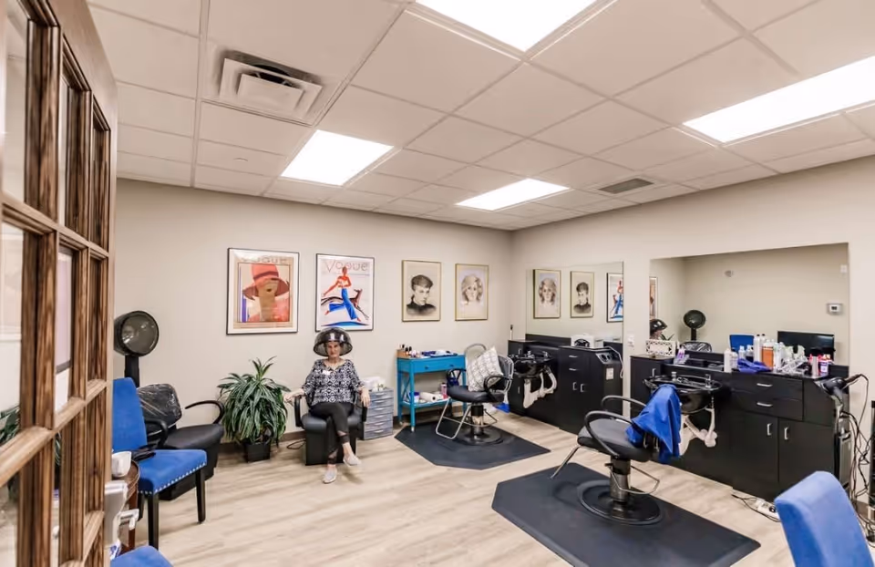 Interior of a salon room in a retirement community with a woman sitting under a hair dryer. The room has salon chairs, hair washing stations, a plant, and framed artwork on the walls.