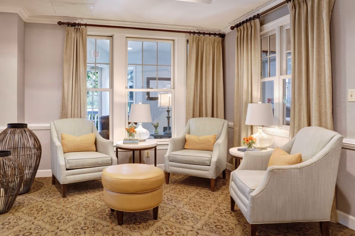 A cozy sitting area with three light-colored armchairs each with a yellow cushion, two round side tables with lamps and small flower arrangements, a round tan ottoman in the center, beige curtains on windows, and decorative wicker baskets on the side, all set on a patterned carpet.