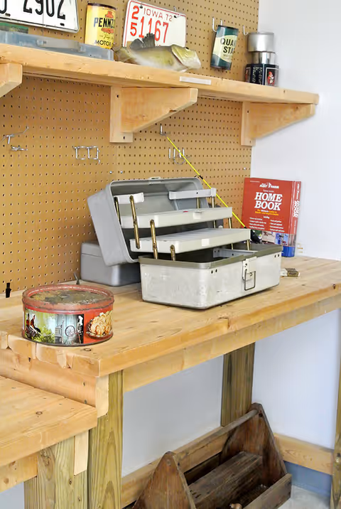 Wooden workbench against a pegboard holding an open metal tackle box, fishing gear, cans, and a 'Home Book' on a shelf.