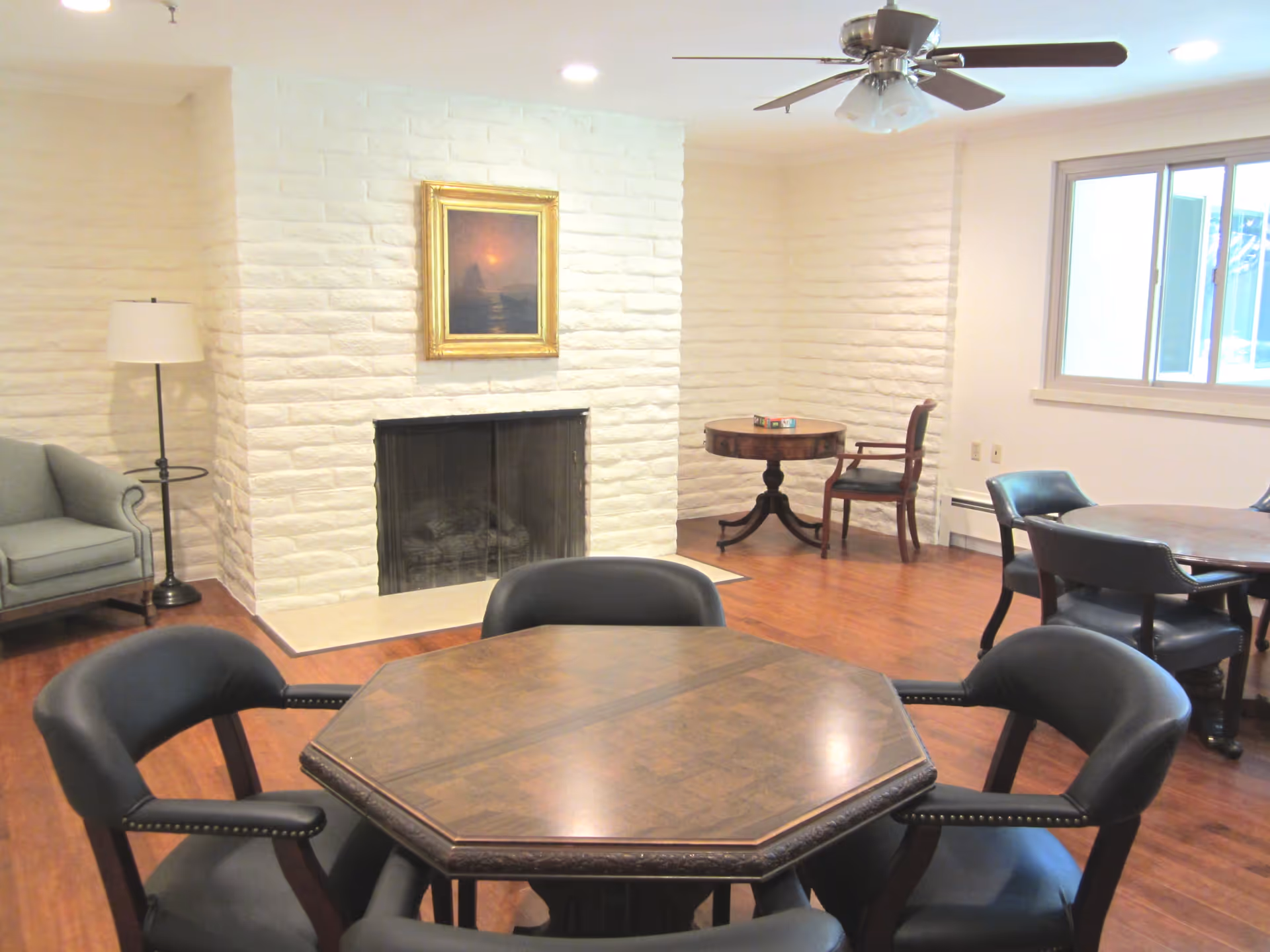 A bright common room with an octagonal wood table and chairs in the foreground, a white-brick fireplace, and additional seating and tables.