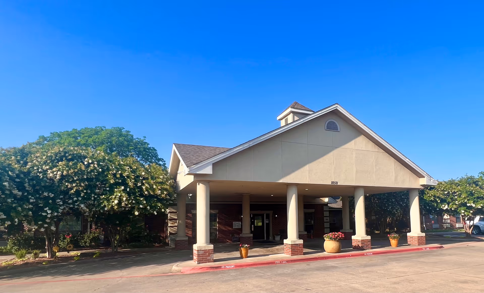 Front exterior view of Millbrook Healthcare and Rehabilitation Center building with a covered entrance supported by columns, surrounded by trees and plants under a clear blue sky.