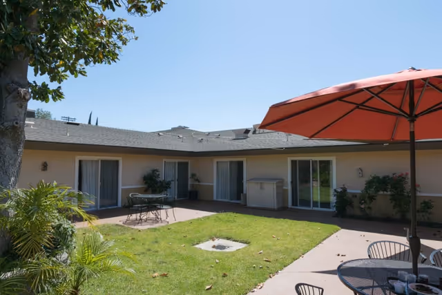 Sunny courtyard with grass, patio tables and a red umbrella surrounded by single-story nursing facility rooms with sliding glass doors.