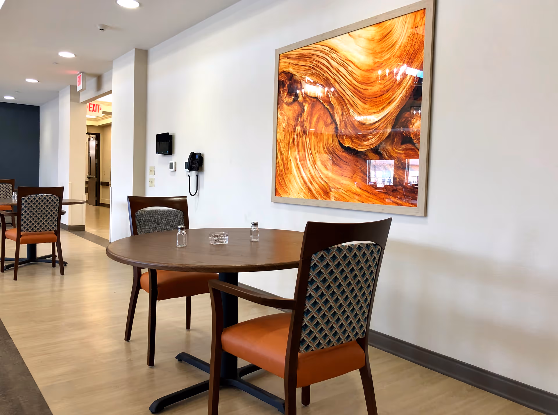 Interior view of a dining area with round wooden tables and chairs with patterned backs and orange seats. On the wall is a large framed artwork featuring a close-up of wood grain with rich brown and orange tones. The floor is light-colored wood, and there is a hallway visible in the background with an exit sign.
