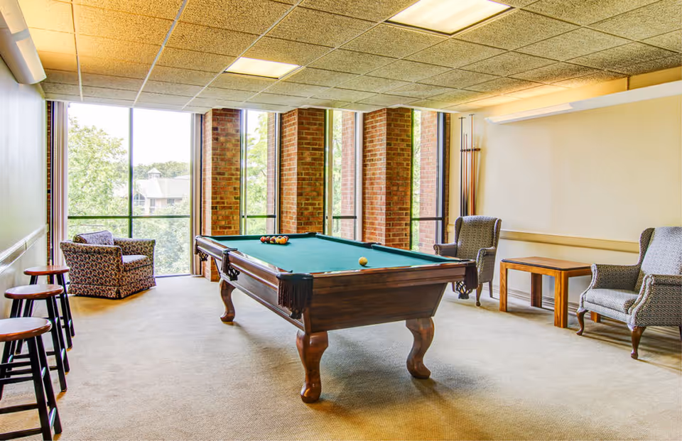 A bright room with a pool table in the center, surrounded by several chairs and stools. Large windows with brick columns allow natural light to fill the space, and a set of pool cues is mounted on the wall.