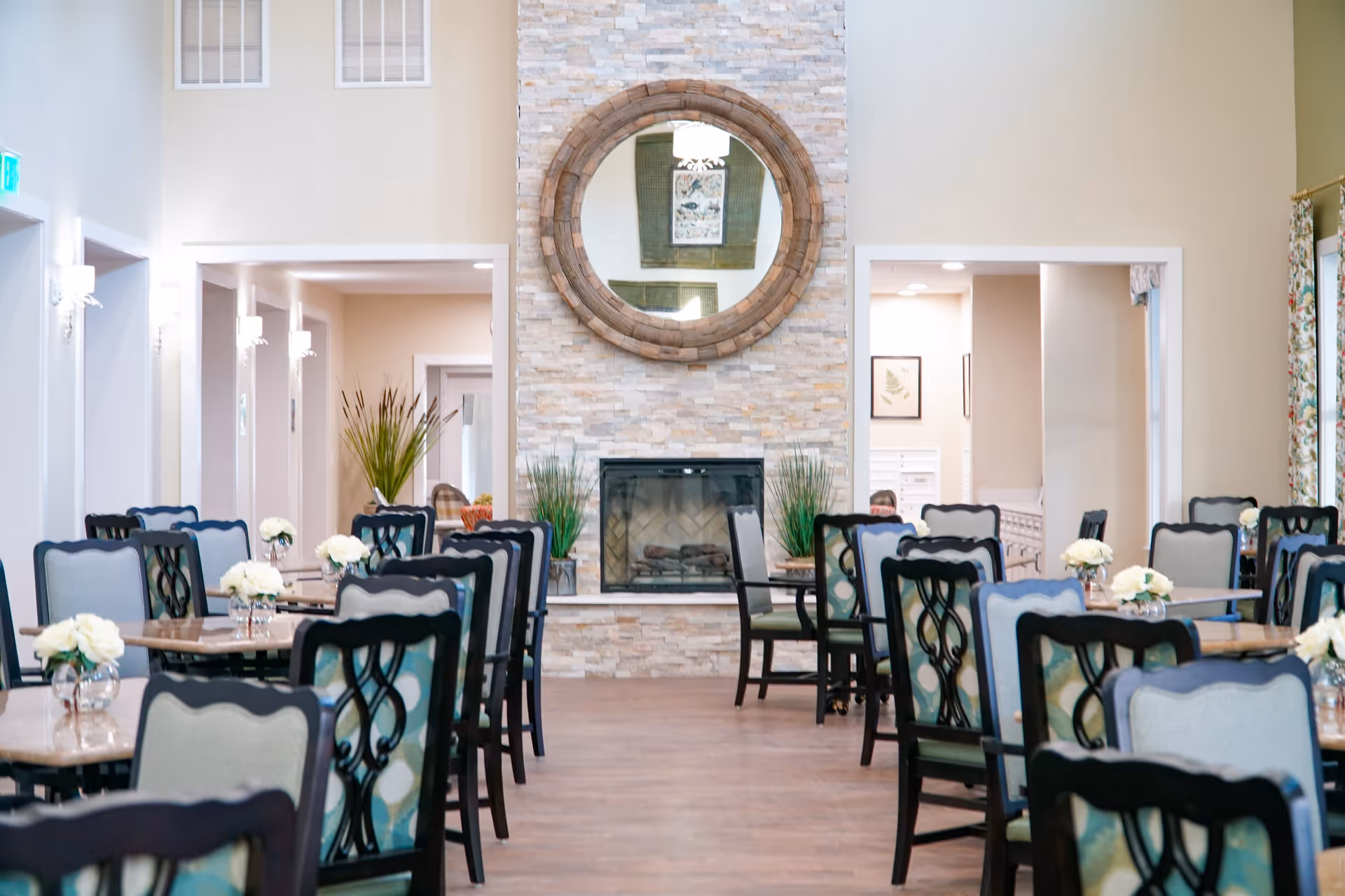 A bright and elegant dining room with multiple tables and chairs arranged neatly. Each table has a small vase with white flowers. The room features a stone fireplace with a large round mirror above it. The walls are painted in light colors, and there are decorative plants and framed artwork visible in the background.