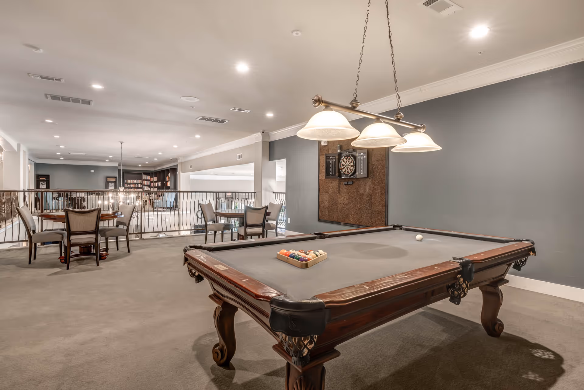A recreational room with a pool table in the foreground, a dartboard mounted on the wall, and several tables with chairs in the background. The room has a gray carpet, gray walls, and recessed ceiling lights.