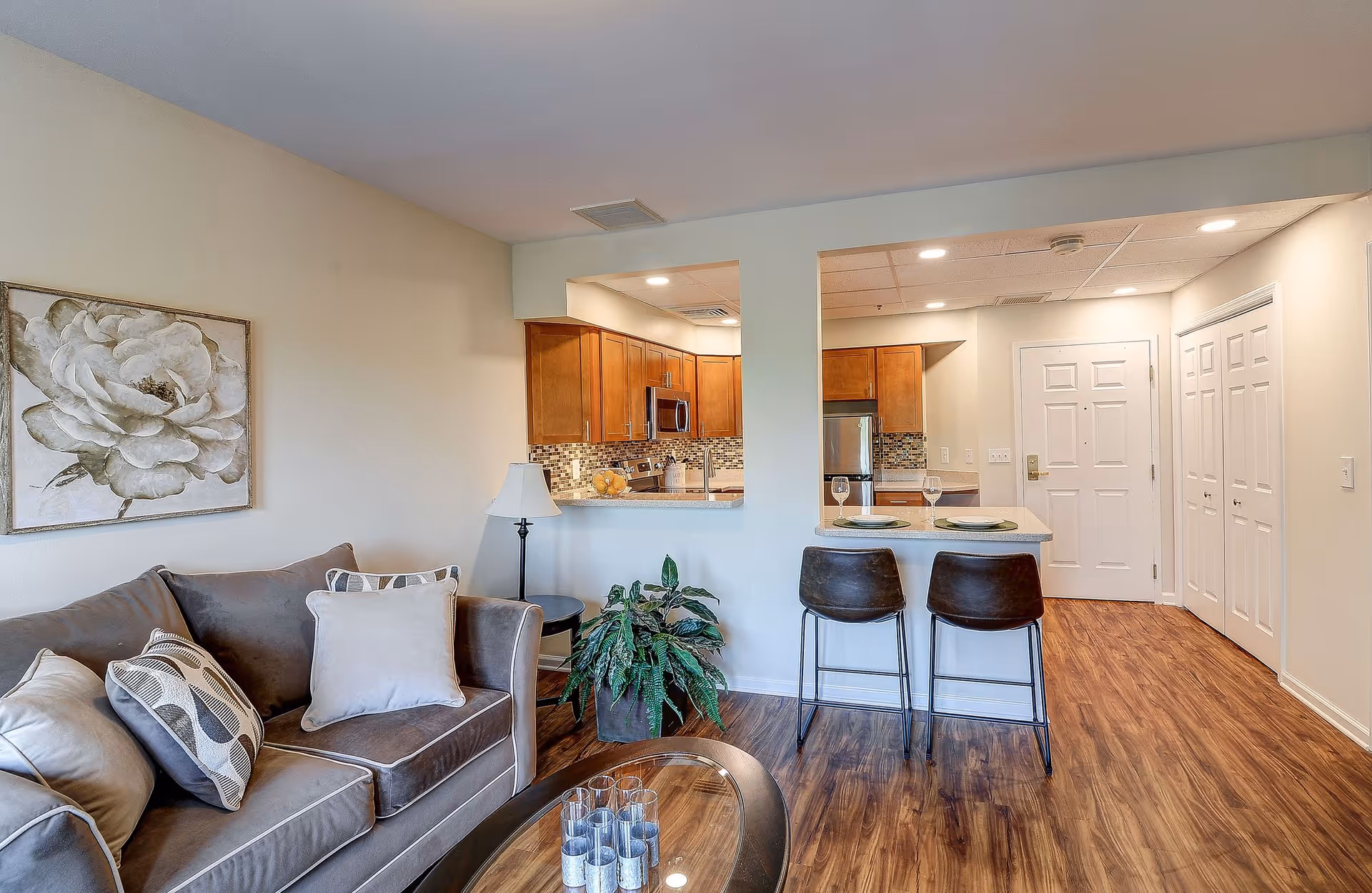 A modern living area with a gray sofa adorned with multiple pillows, a glass coffee table with candles, a floor lamp, and a large floral painting on the wall. The living room opens into a kitchen with wooden cabinets, stainless steel appliances, and a breakfast bar with two stools set with plates and wine glasses. The floor is wood, and the walls are painted light beige.