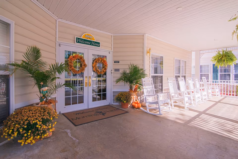 Covered porch entrance of a senior living facility with white double doors decorated with autumn wreaths, a welcome mat that reads 'Morning Pointe', several white rocking chairs lined up along the porch railing, potted plants including ferns and flowers, and pumpkins placed near the door.