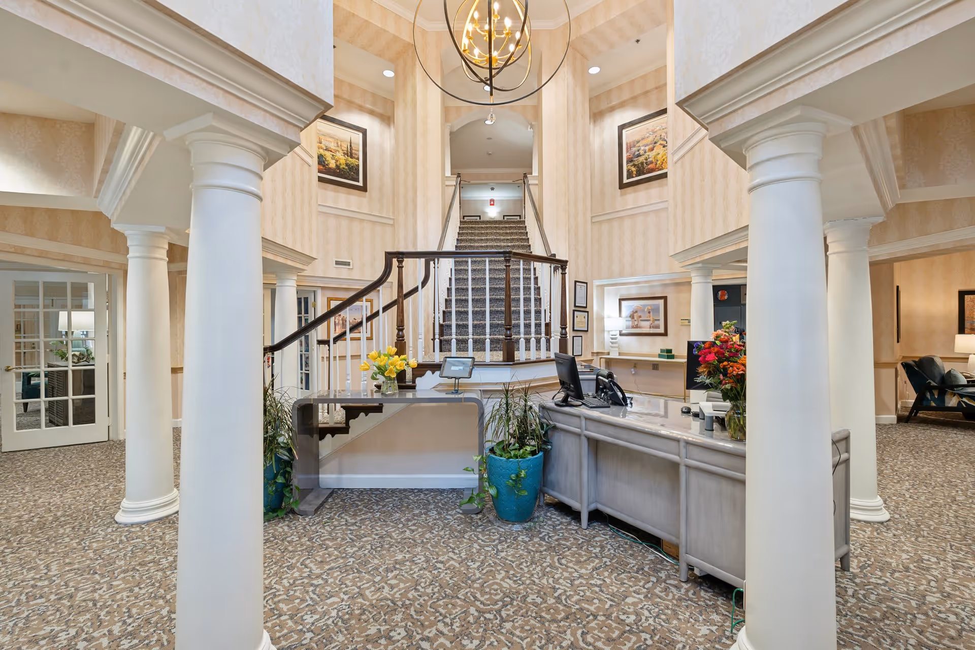 Interior view of a senior living facility lobby with a reception desk, decorative columns, a staircase in the background, framed artwork on the walls, and floral arrangements on the desk.
