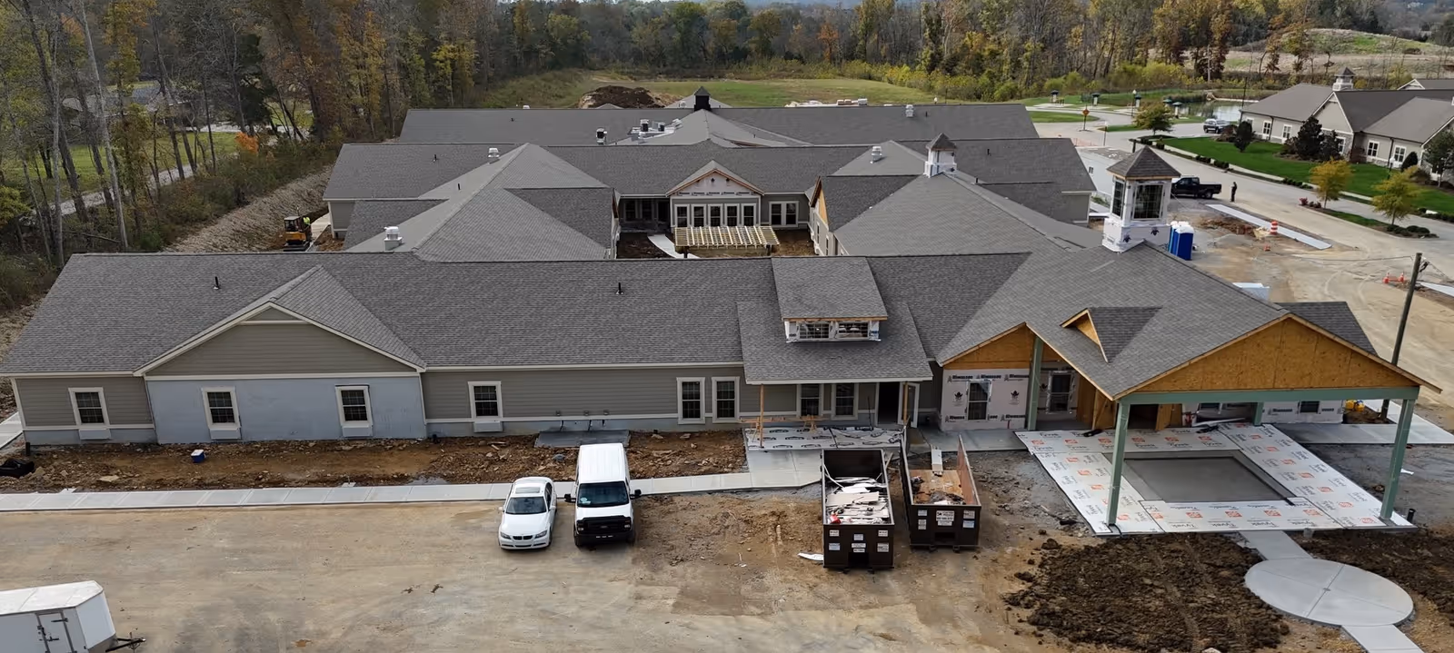 Aerial view of a large building under construction with multiple roof sections and a covered entrance area. Two vehicles are parked in front, and construction materials and dumpsters are visible. The surrounding area includes trees and other buildings in the distance.