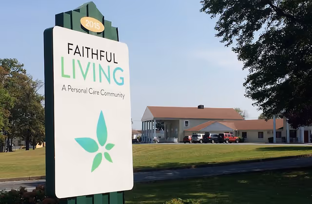 Outdoor view of Faithful Living, a personal care community, showing a large sign with the facility name and logo in front of a building with a parking area and several vehicles under a clear sky.