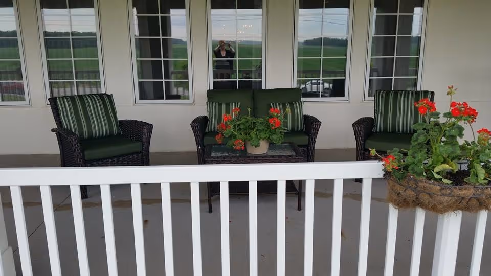 Outdoor patio area with three dark wicker chairs featuring green cushions and striped pillows, a small glass-top table with a potted plant with red flowers, and a white railing in the foreground. There are windows in the background reflecting the outdoor scenery and a person taking the photo.