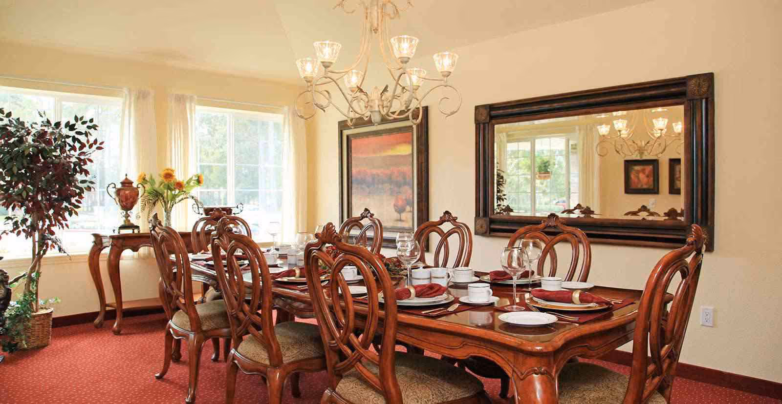 Formal dining room with an ornate wooden table set with dishes and glassware, matching chairs, a chandelier, large mirror, and windows letting in daylight.