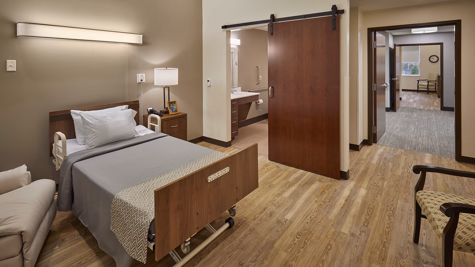 A clean, furnished senior bedroom with a hospital-style bed, bedside table and lamp, a sliding wooden barn door to a bathroom, and chairs visible in the adjoining hallway.