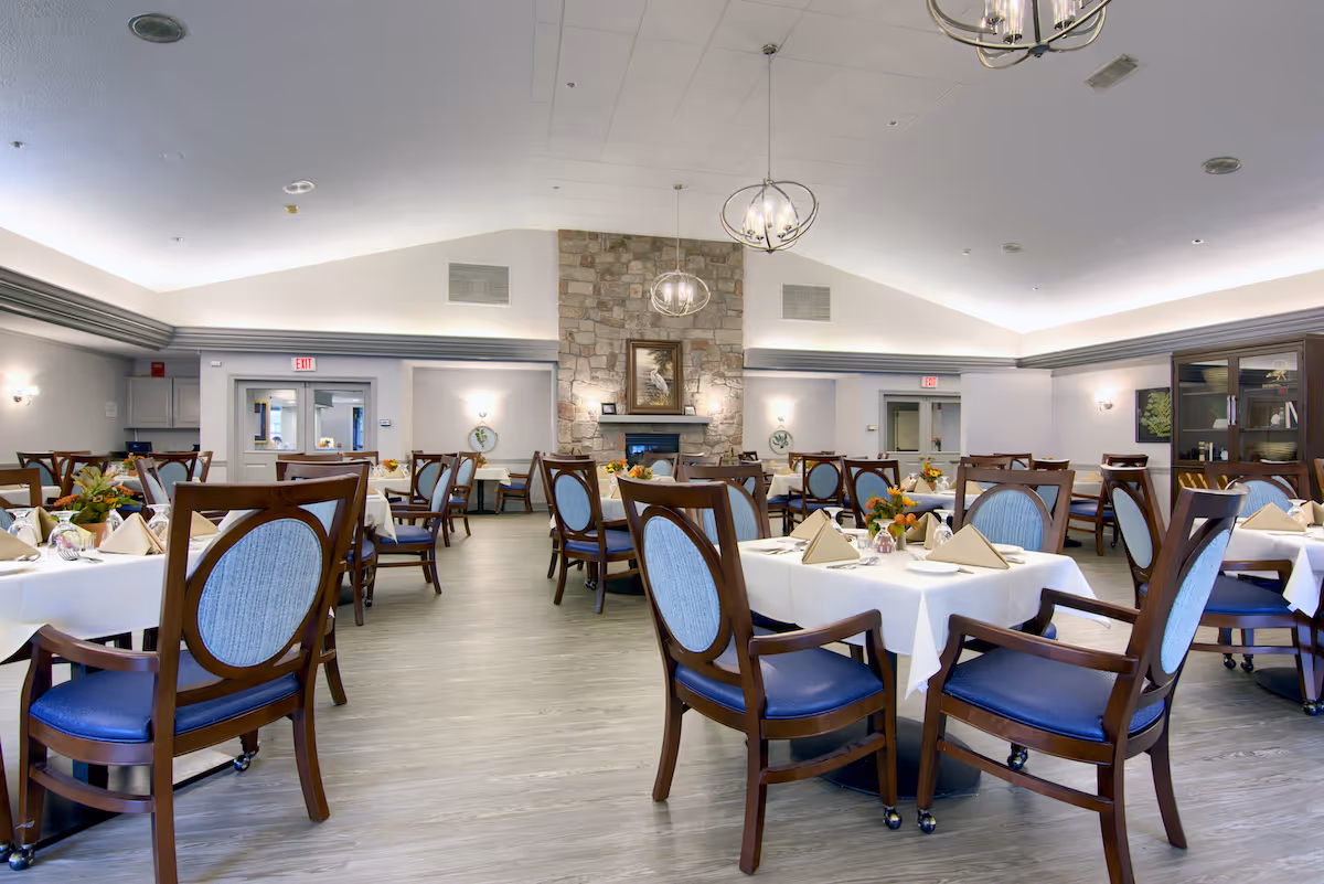 A spacious senior living community dining room with multiple tables covered in white tablecloths, each set with napkins, plates, and floral centerpieces. The room features wooden chairs with blue cushions, a stone fireplace with a framed picture above it, and modern hanging light fixtures. The walls are light-colored, and there are exit signs above doors in the background.
