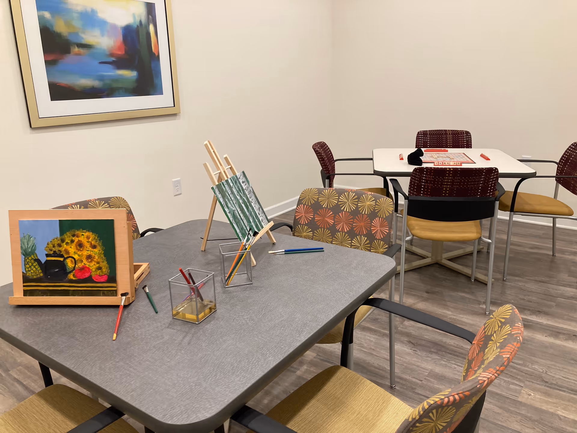 A room with two tables and several chairs. One table has painting supplies including paintbrushes, two small paintings on easels, and containers holding brushes. The other table has a board game set up. The room has light-colored walls, wood flooring, and a colorful abstract painting hanging on the wall.