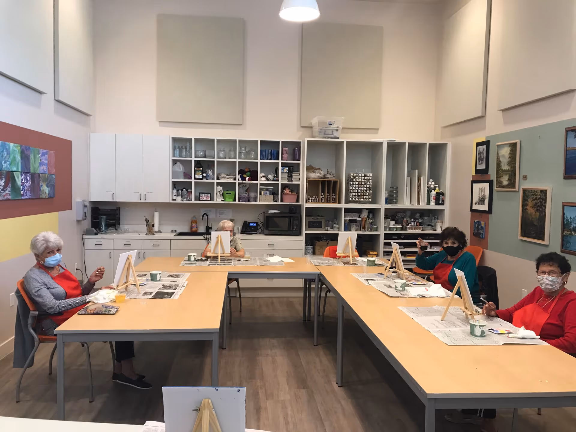 Several older adults wearing masks sit at tables in a well-lit art room painting at small easels.