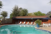 Outdoor swimming pool with lounge chairs and tables in front of a low pool house and trees.