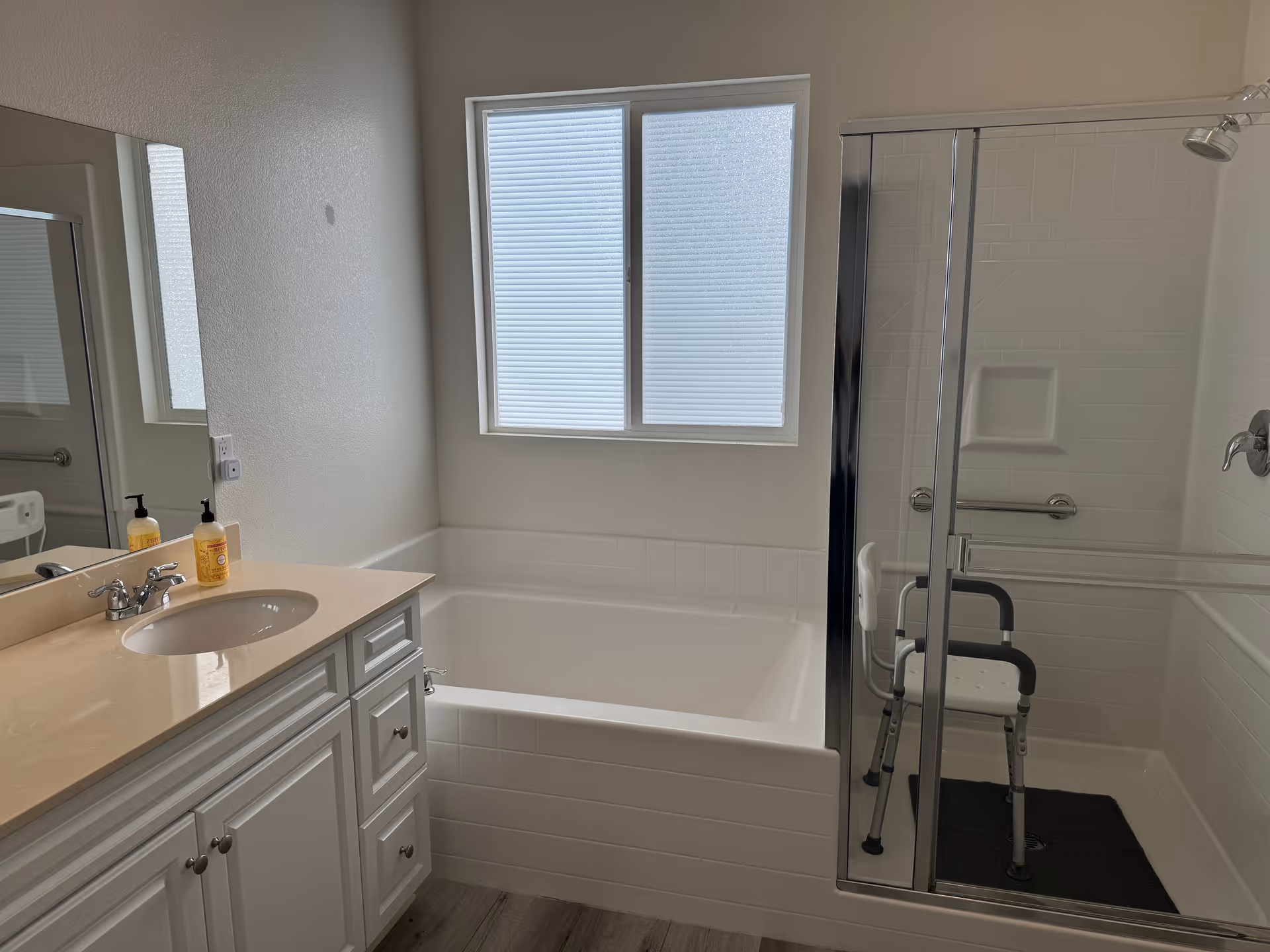 A clean bathroom with a white vanity and sink on the left, a large bathtub beneath a frosted window in the center, and a glass-enclosed shower with a shower chair and grab bars on the right.
