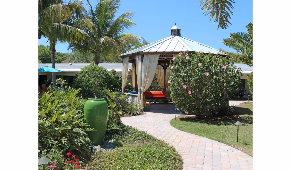 A sunny outdoor garden area with a paved walkway leading to a gazebo with curtains and colorful seating. The garden features lush green plants, palm trees, a large green ceramic vase, and a flowering bush with pink flowers. A building with a gray roof is visible in the background under a clear blue sky.